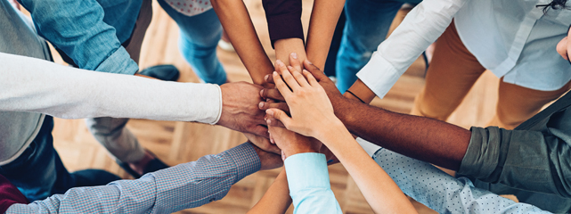 eight people from differnt racial and ethnic groups' arms pointed to a central point with their hands are stacked upon eachother as a team.