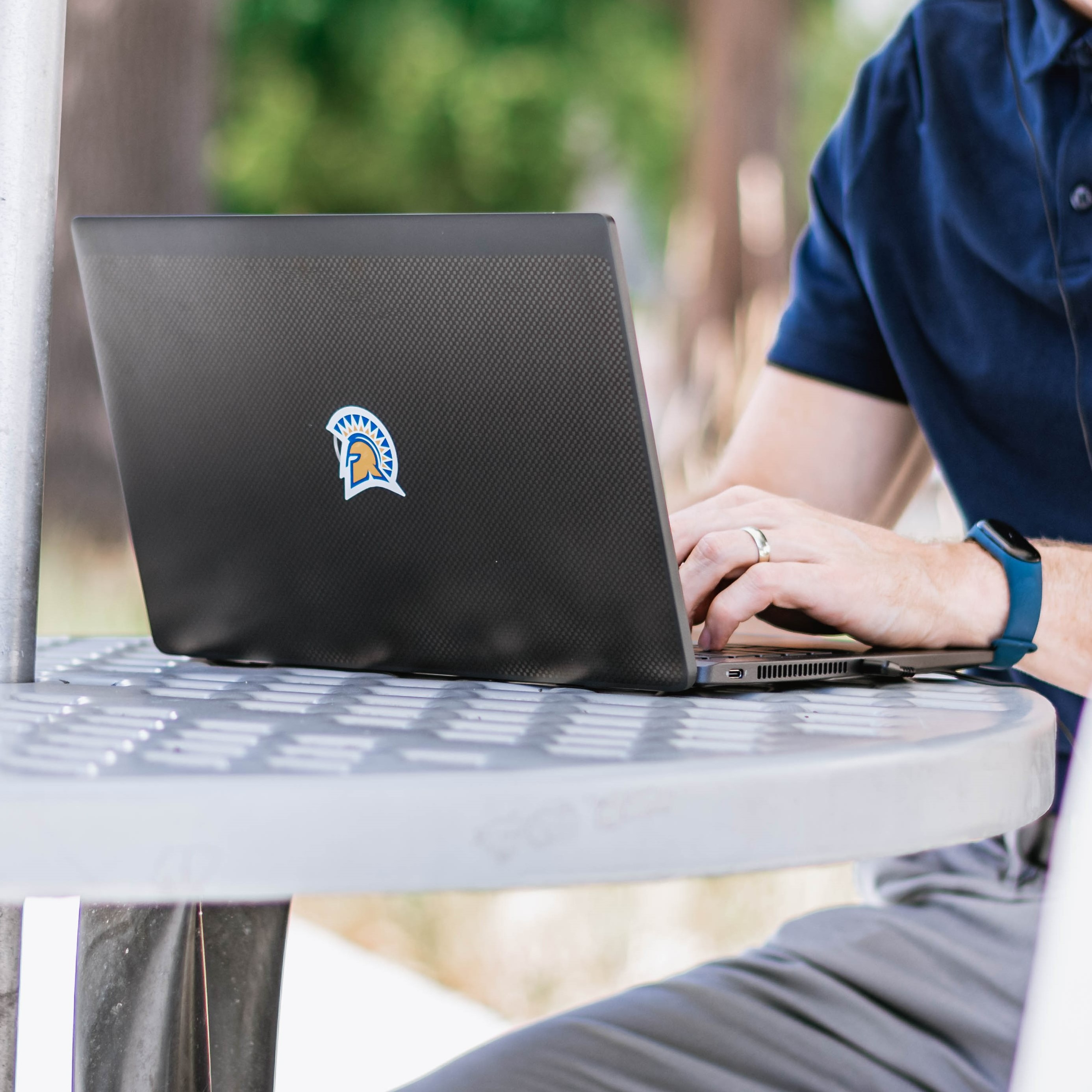 A student wearing a gray baseball cap ponders with his hand to his mouth while they stare at their laptop